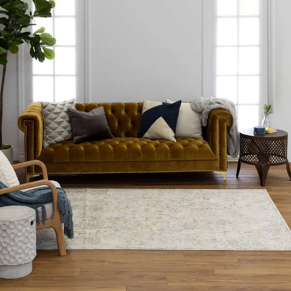 Light-colored area rug with subtle pattern placed in a modern living room setting with velvet tufted sofa and decorative pillows