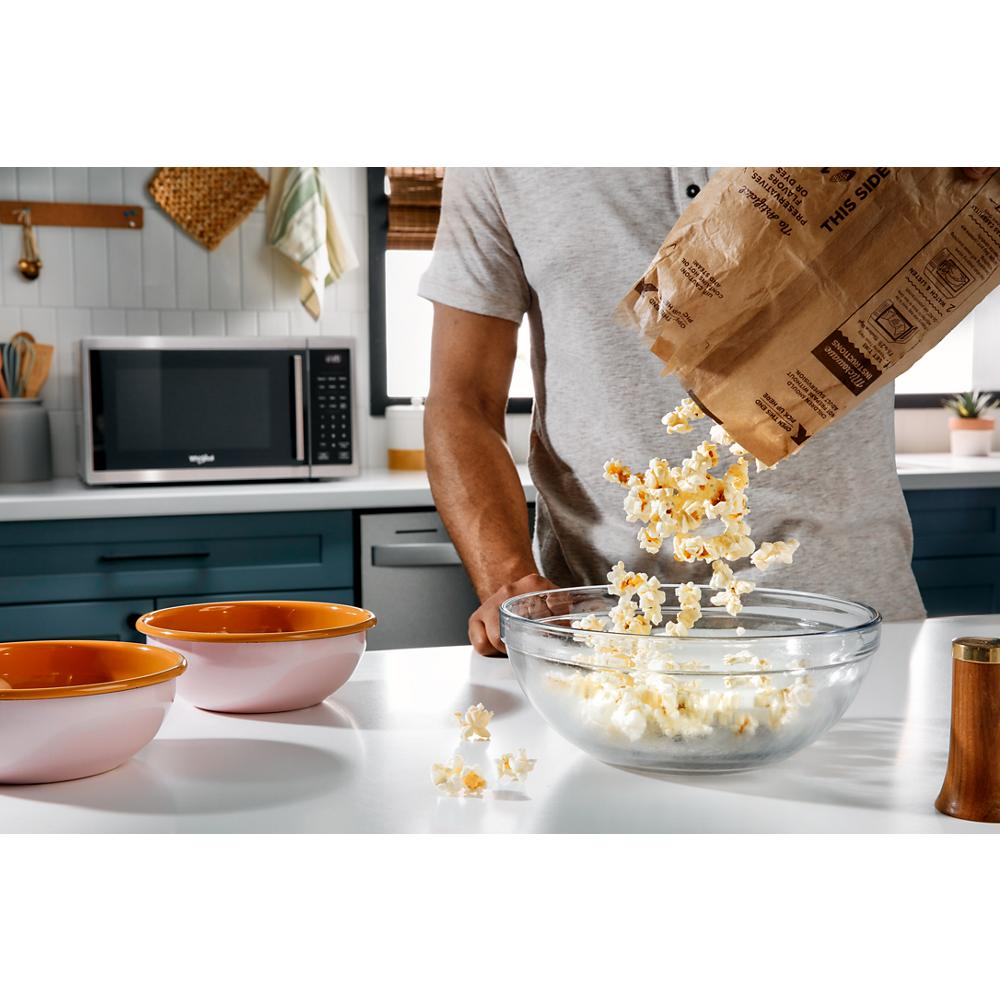 Person pouring popcorn from a paper bag into a glass bowl on a kitchen counter with black microwave in background