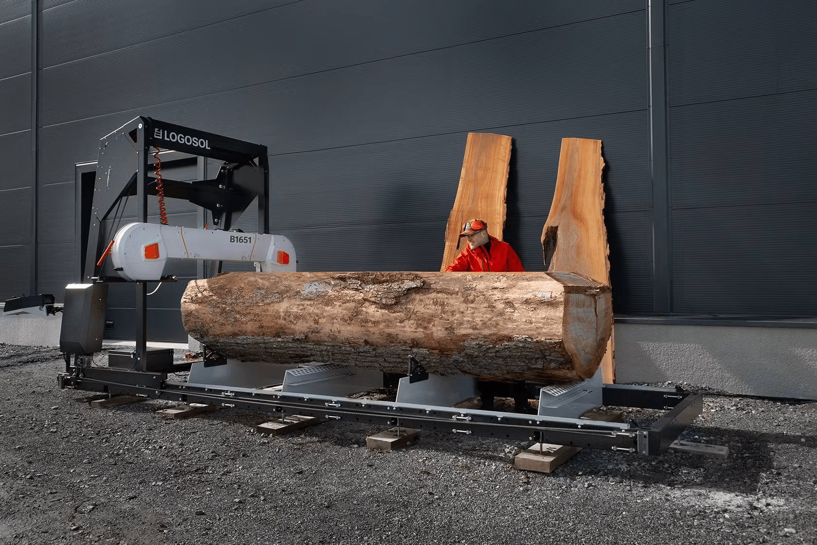Portable band sawmill with 27HP gasoline engine cutting a large log outdoors against a dark industrial wall