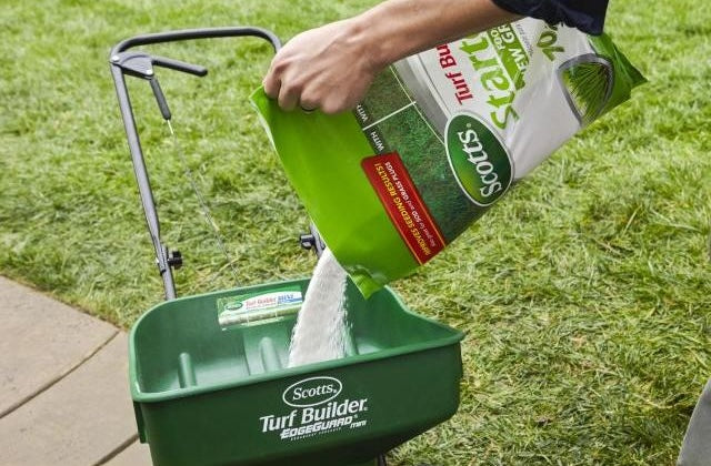 Pouring Scotts starter fertilizer into a green Turf Builder spreader on a grassy lawn in lawn care and plant fertilizer use