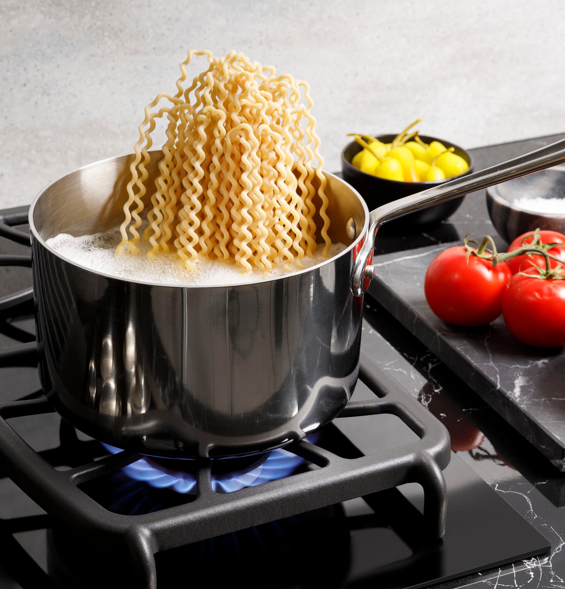 Stainless steel pot boiling wavy pasta on gas cooktop burner with tomatoes and peppers nearby in kitchenware setting