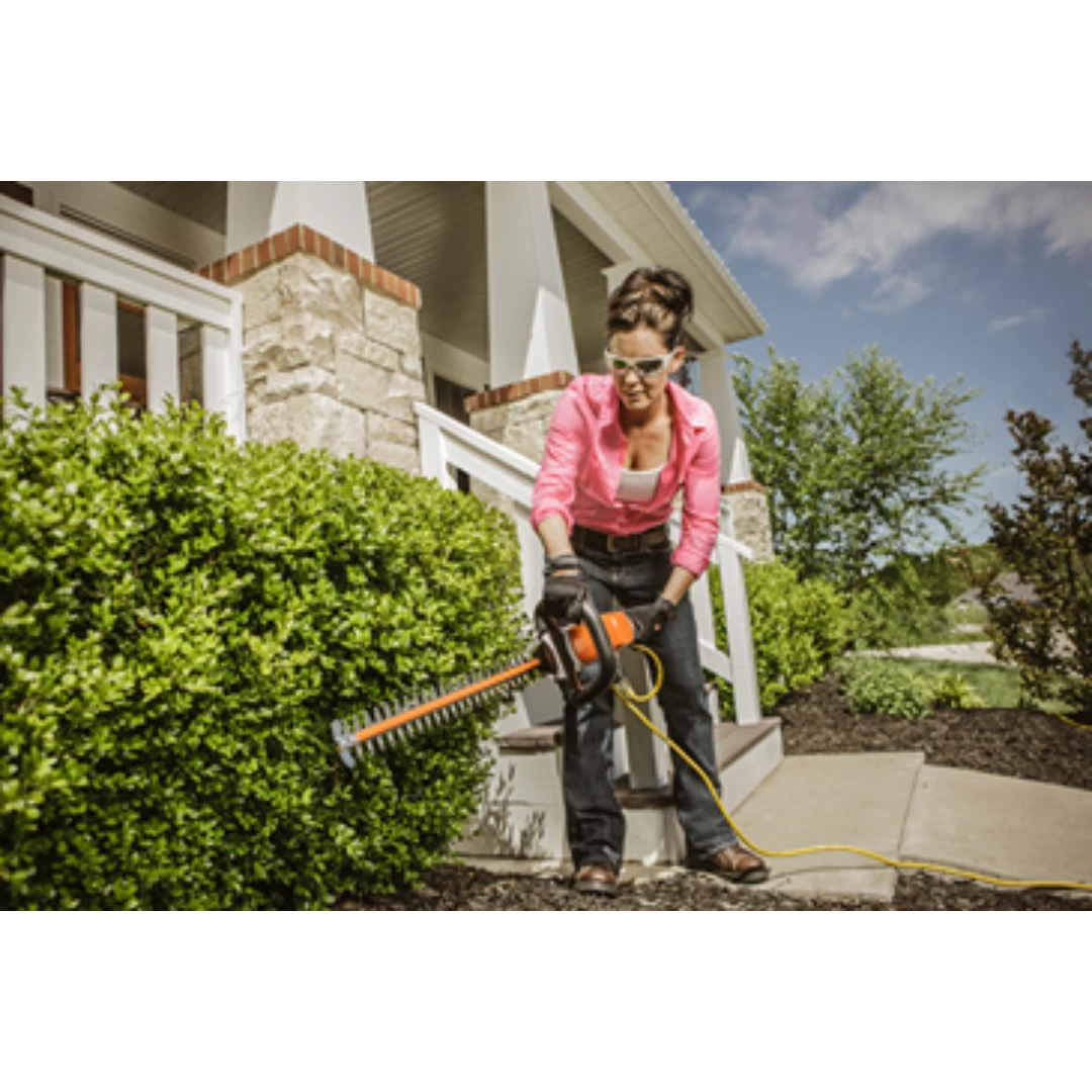 Woman wearing protective glasses using orange and black electric hedge trimmer to shape green shrubs outdoors near home