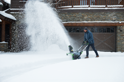 Man using a cordless snow blower with steel auger clearing heavy snow outside a home in a winter setting