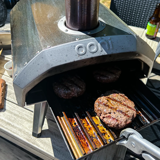 Compact outdoor pizza oven with grilled burgers cooking on a metal grate, ideal for outdoor living and grilling accessories
