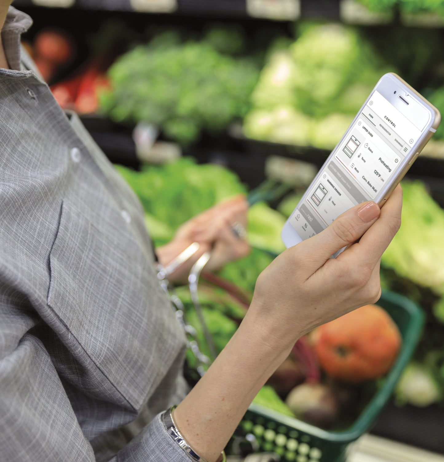 Person holding smartphone with oven control app open, standing near fresh produce in grocery store setting
