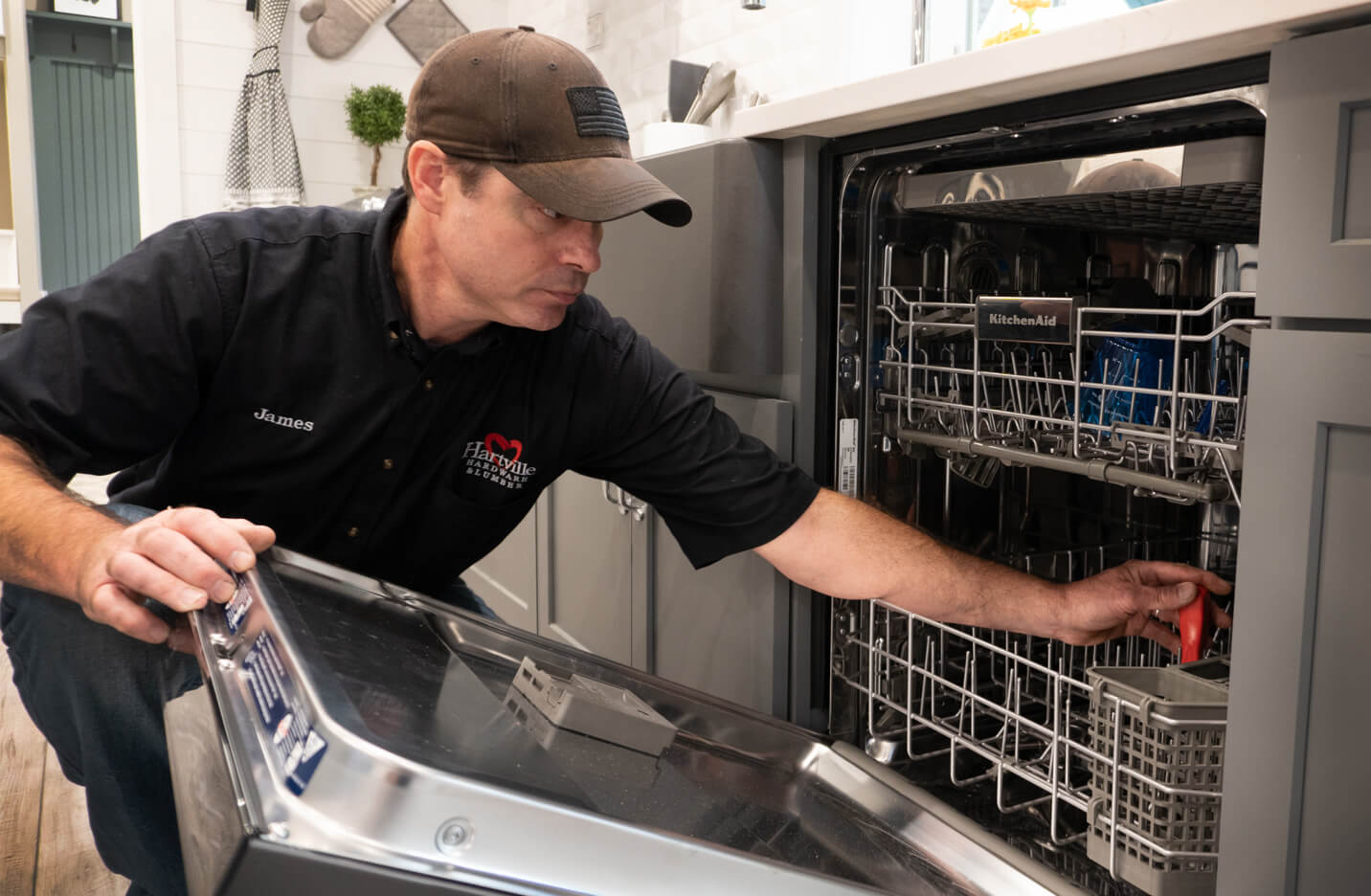 Man wearing a black shirt and cap loading or adjusting dishes inside a stainless steel dishwasher in a modern kitchen setting.