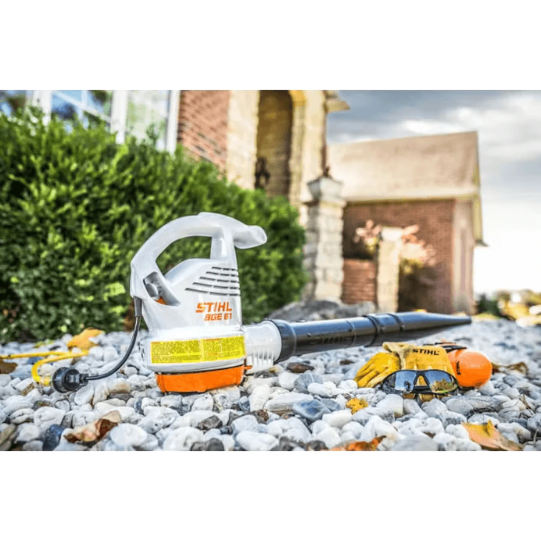 White and orange electric handheld blower with safety gloves and goggles on gravel in outdoor home setting