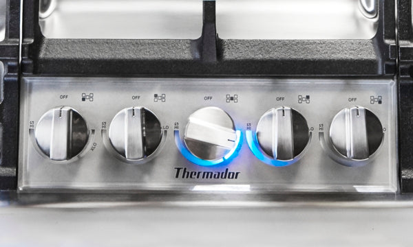 Close-up of stainless steel cooktop control panel with five knobs and illuminated blue flames from Thermador collection