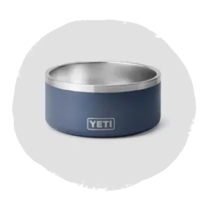 Small round pet bowl with a stainless steel interior and a matte blue exterior featuring a simple logo on the front, shown on a neutral background.