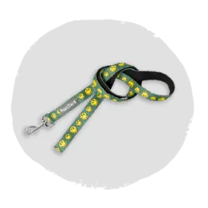 Green fabric dog leash printed with yellow smiley faces, featuring a black padded handle and a metal clip on a light gray background.