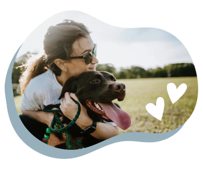 Woman wearing sunglasses smiles while holding a happy black dog with its tongue out in a sunny outdoor field with heart graphic accents.