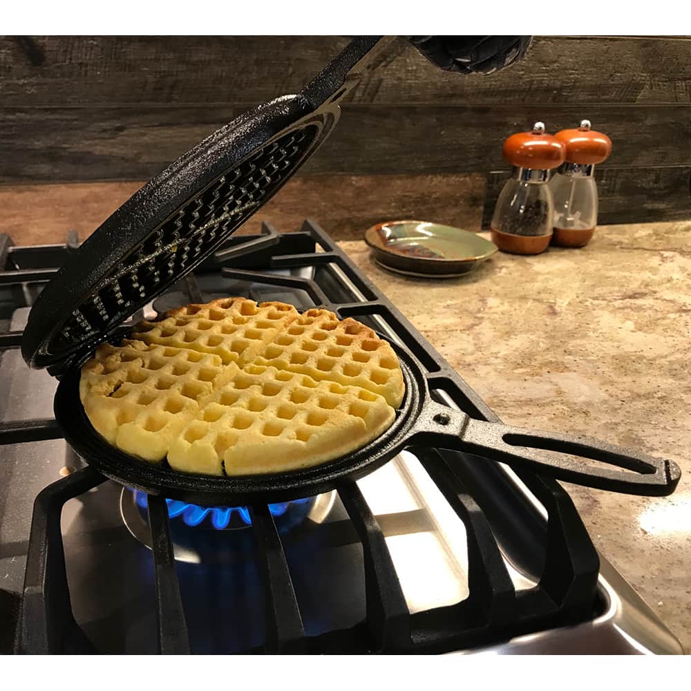 Black cast iron stovetop waffle maker with long handle cooking waffle over blue gas flame on stove burner