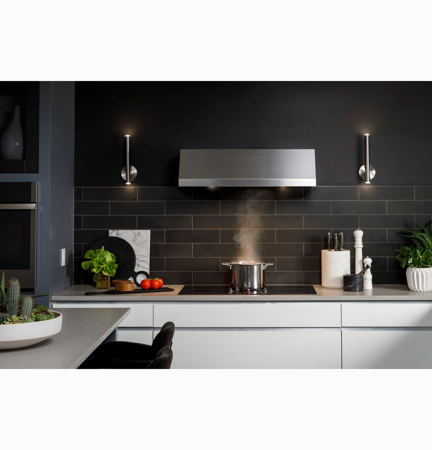 Modern stainless steel kitchen range hood above stovetop with cooking pot, surrounded by kitchenware and plants