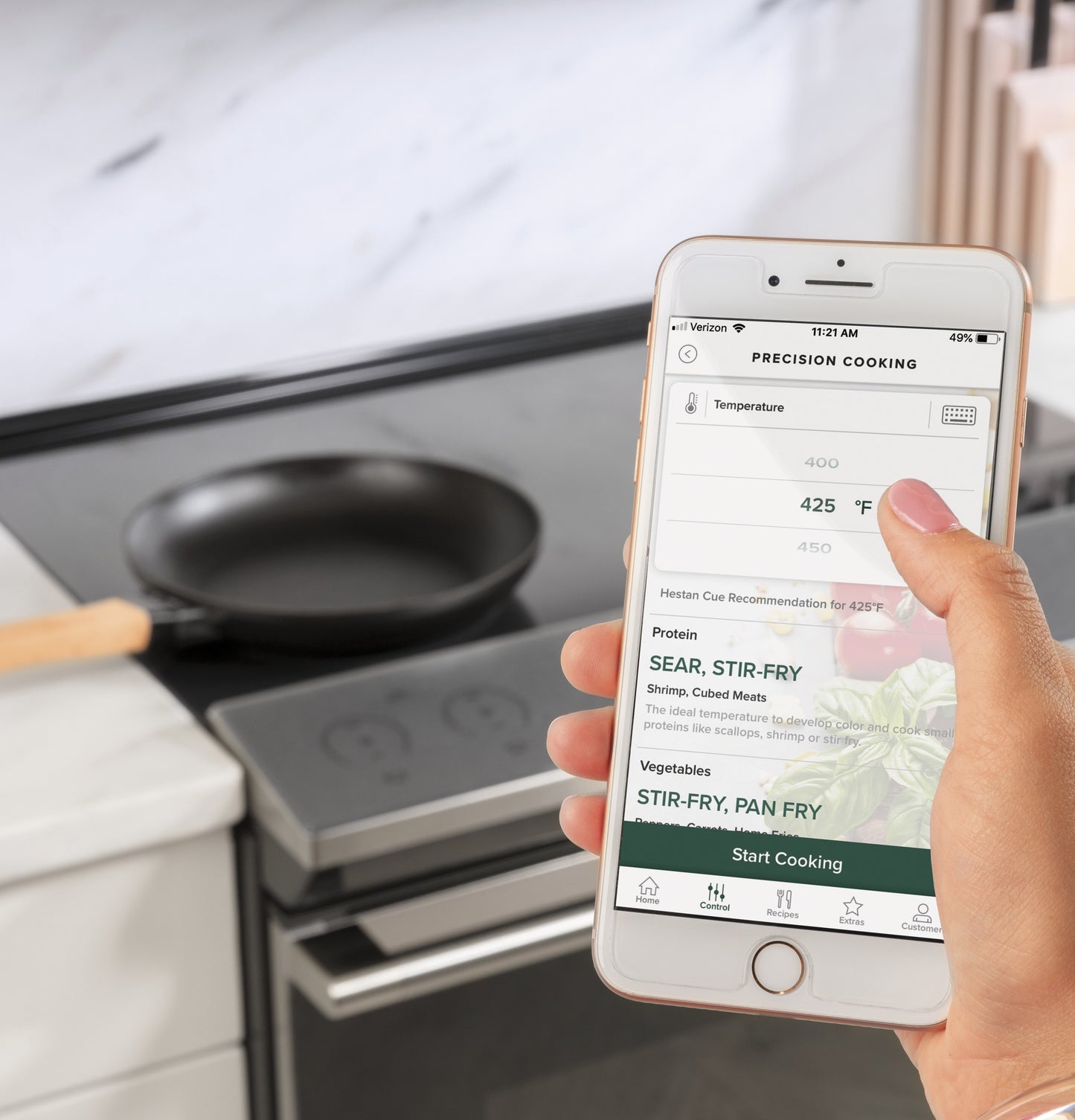 Hand holding smartphone with precision cooking app set to 425°F in kitchen near induction cooktop and skillet