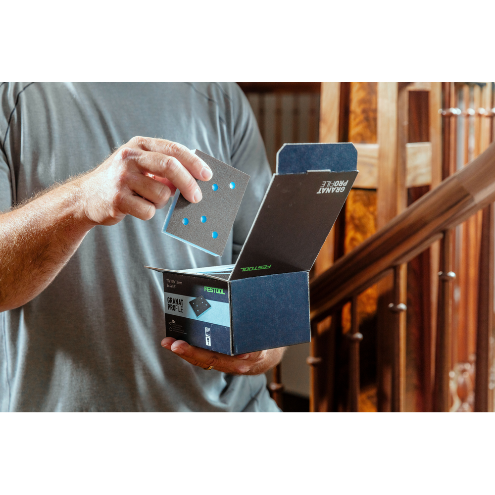 Person holding a blue abrasive sanding pad above an open Festool Grit Abrasives box in a home improvement setting