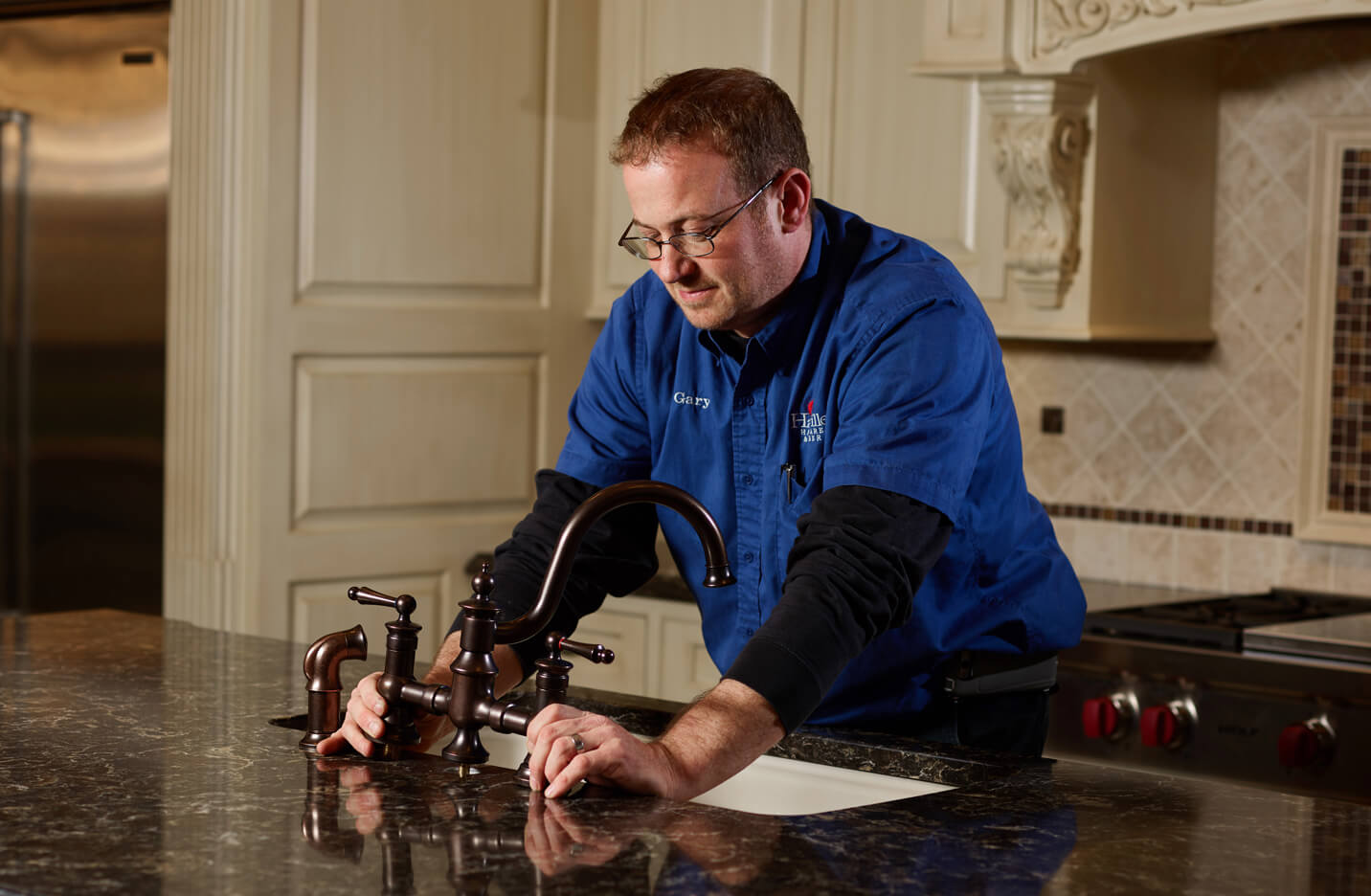 Man in a blue work shirt installing or adjusting a dark bronze kitchen faucet on a polished granite countertop in a traditional kitchen setting.