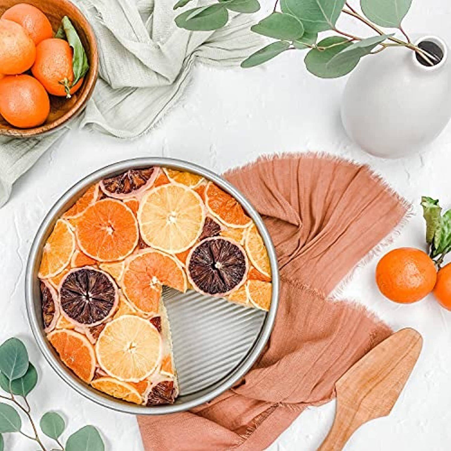 Round metal cake pan filled with citrus slice-topped cake on white surface with cloth and green leaves nearby