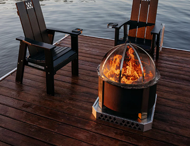 Outdoor fire pit with protective screen and glowing flames on wooden deck between two black Adirondack chairs
