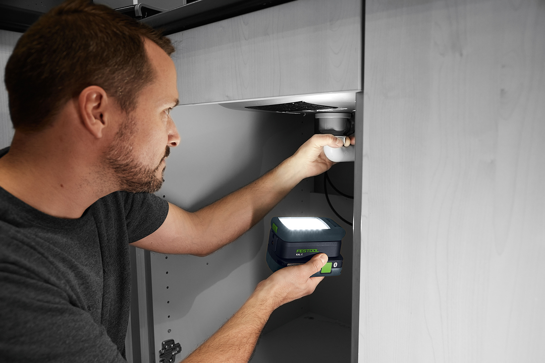 Man using cordless Festool Syslite task light to illuminate plumbing inside a cabinet for home improvement tools
