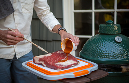 Person pouring sauce onto raw beef brisket on an orange and gray prep tub near a green ceramic grill in outdoor living setting