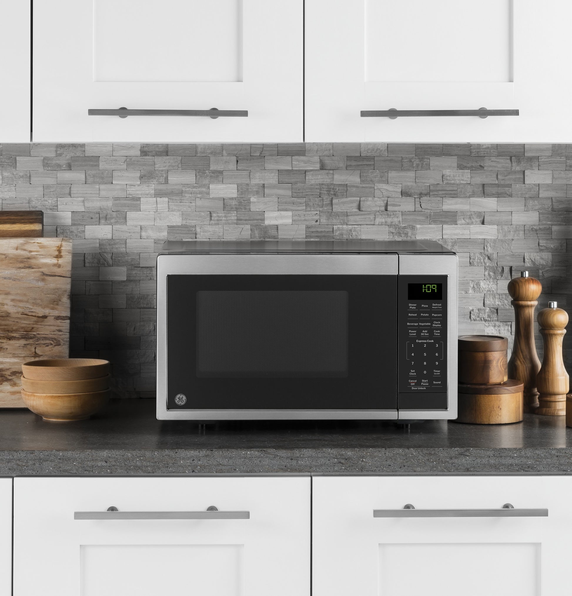 Compact stainless steel microwave on dark countertop with digital control panel and decorative wooden kitchenware nearby