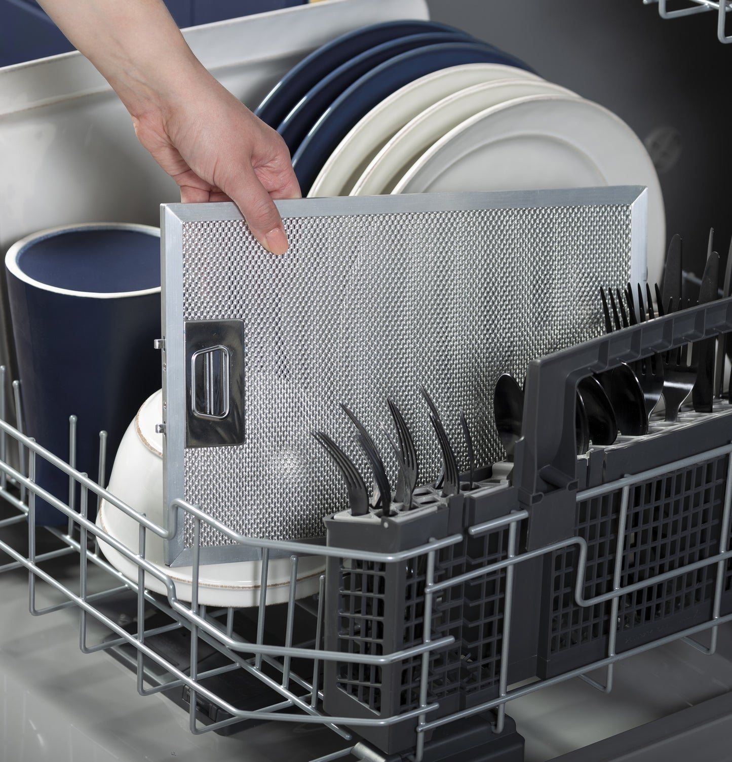 Hand placing a metallic range hood filter into a dishwasher loaded with clean plates and utensils for cleaning appliances