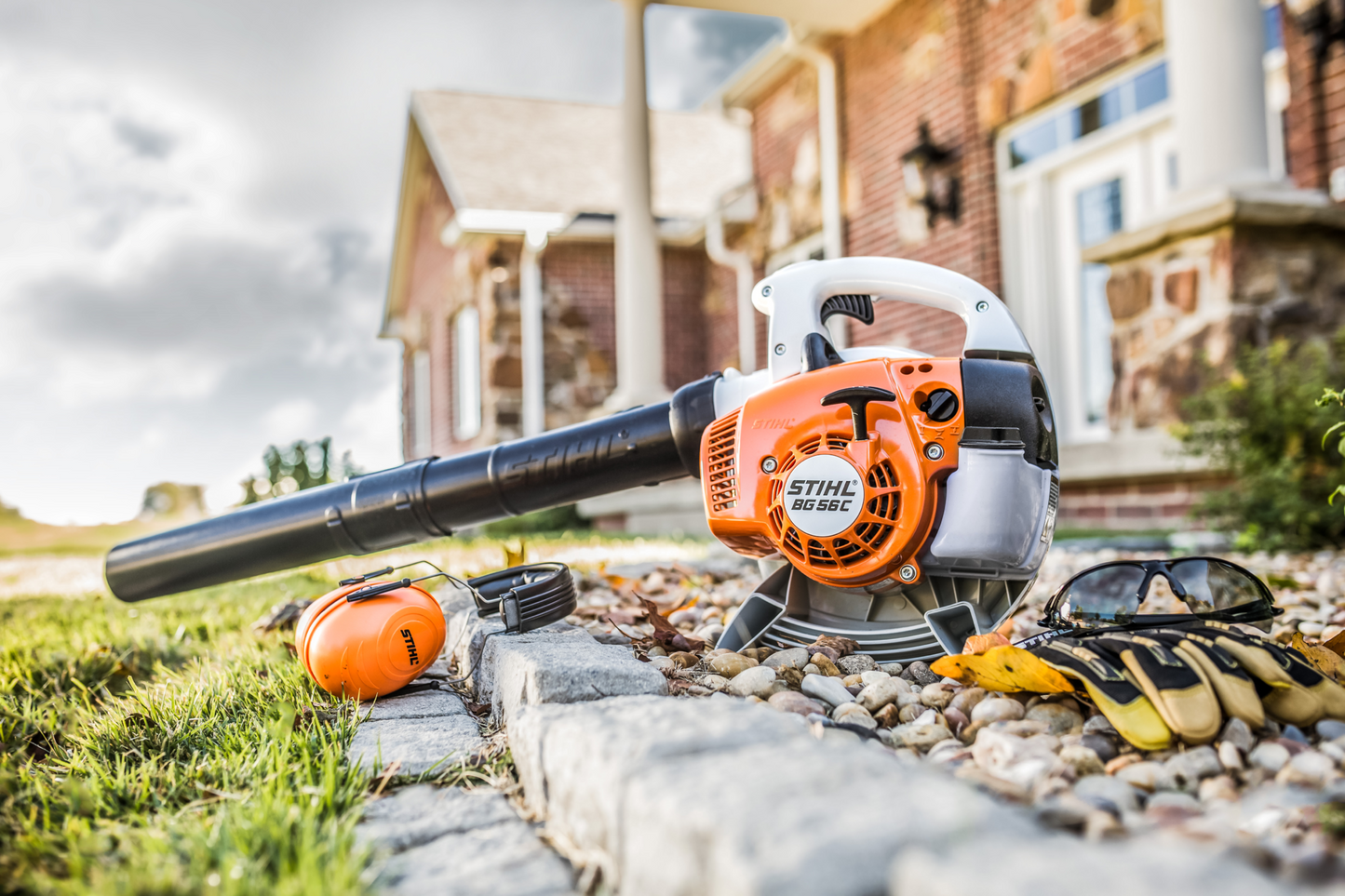 Orange and white handheld blower with black nozzle placed outdoors near gloves and safety earmuffs for yard maintenance tasks