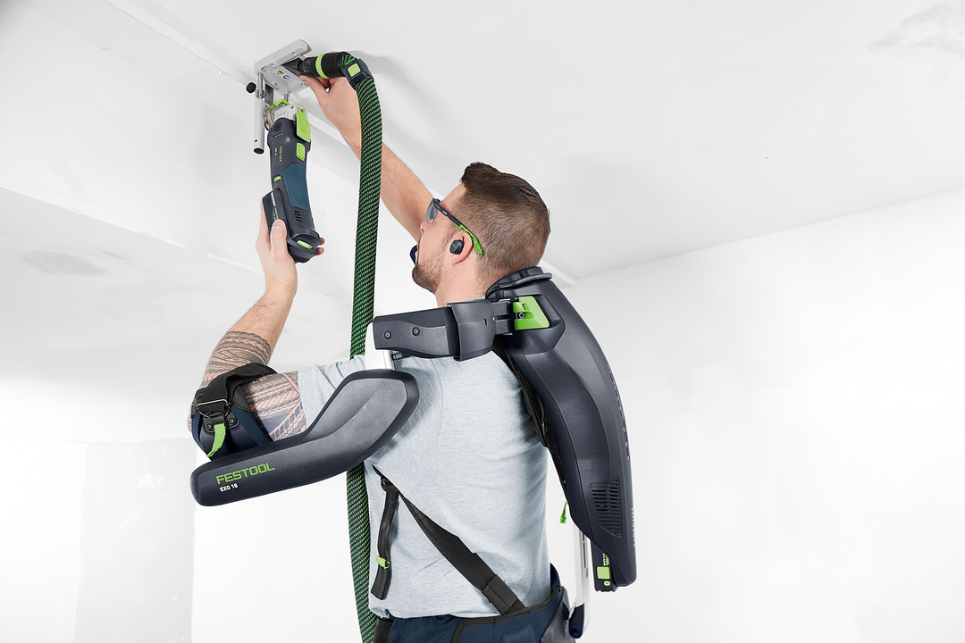 Person wearing Festool ergonomic exoskeleton support system while using power tool for ceiling installation in workshop