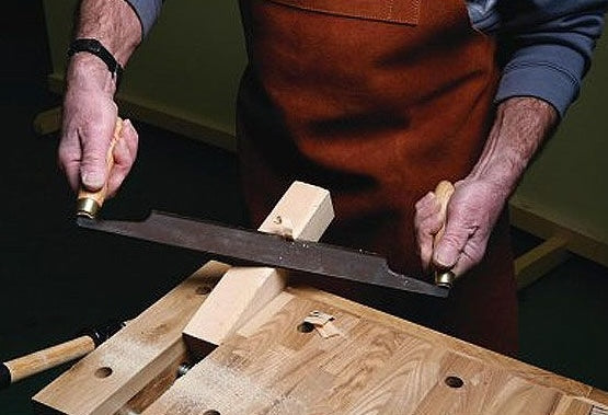 Hands holding a 10-inch bent draw knife shaping wood on a workbench in woodworking tools collection