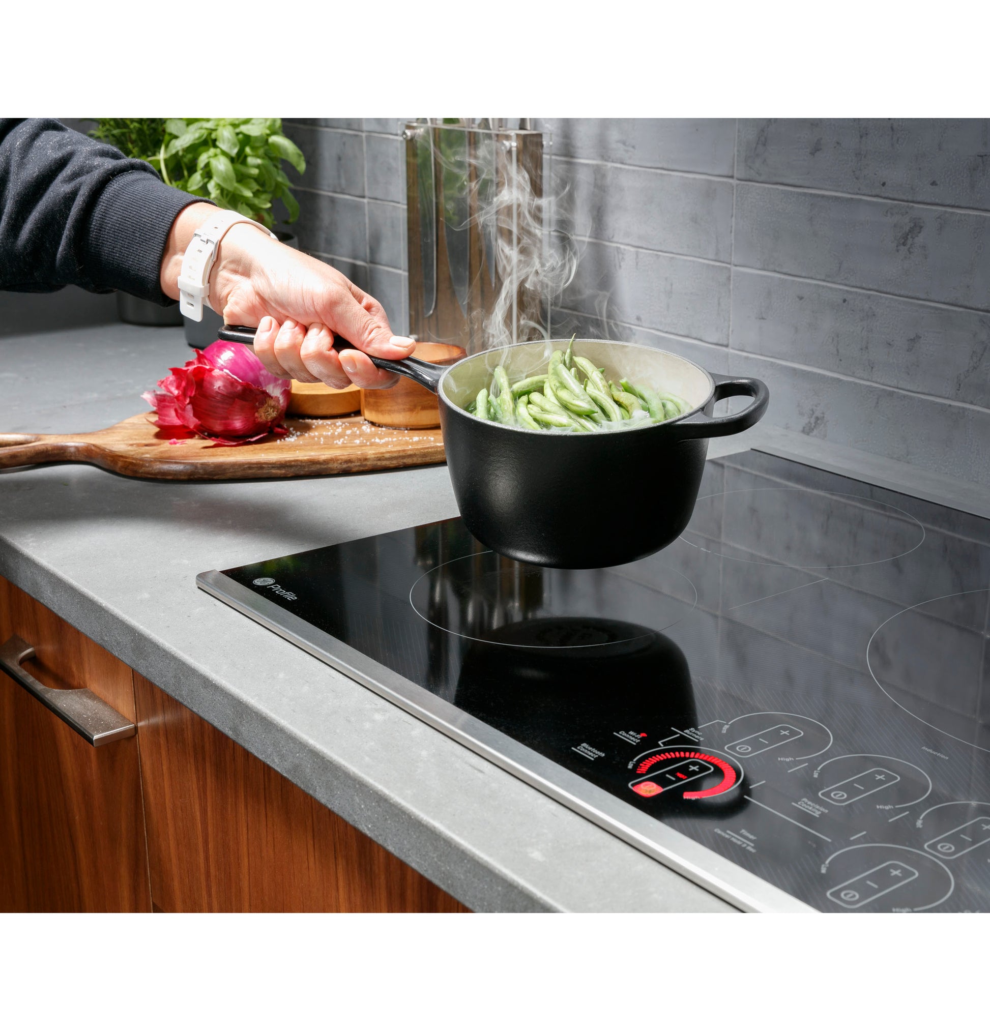 Person holding black pot with steaming green beans above GE Profile glass induction cooktop in kitchen with wood cabinets