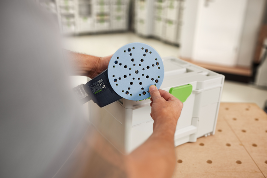 Person attaching blue circular sanding disc with multiple perforations onto power sander in workshop setting tools accessories