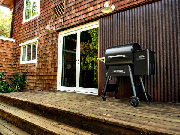 Black pellet grill with digital controls standing on a wooden deck outside a rustic house featuring brown shingle siding and sliding glass doors.