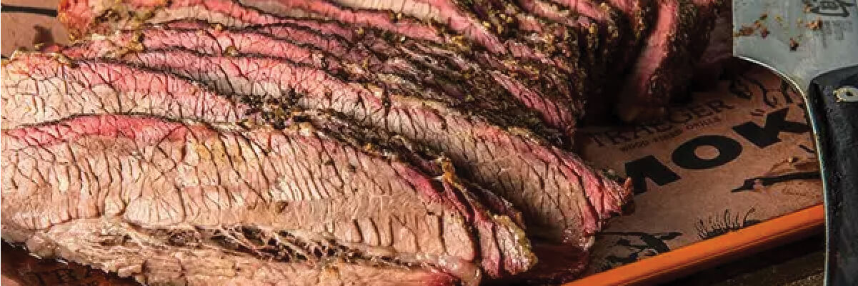 Sliced smoked brisket with a seasoned crust arranged on a branded paper-lined wooden board next to a slicing knife.