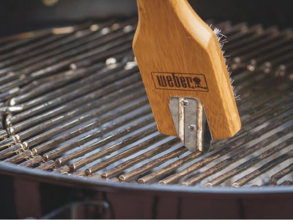 Close-up of a wooden-handled grill brush with metal bristles cleaning a round charcoal grill grate outdoors.
