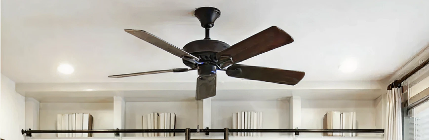 Dark bronze ceiling fan with five wooden blades installed in a bright room featuring built-in shelves lined with decor books and large windows with light curtains.
