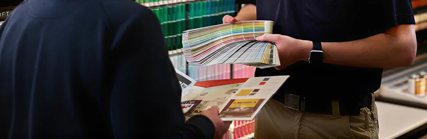 Two people reviewing a color swatch book and printed material inside a store with shelves of paint cans in the background.