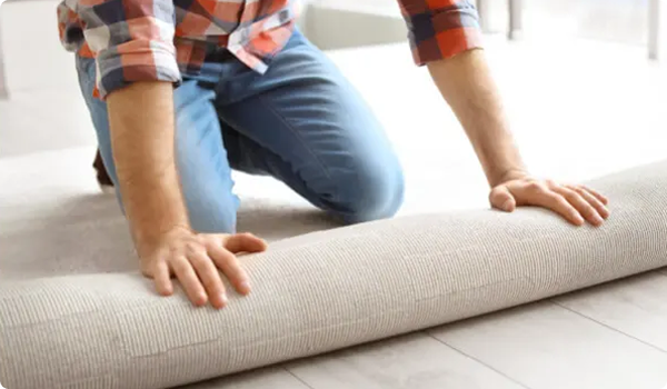 Person wearing a plaid shirt and jeans kneeling on a light wood floor while carefully unrolling a beige textured rug or carpet.