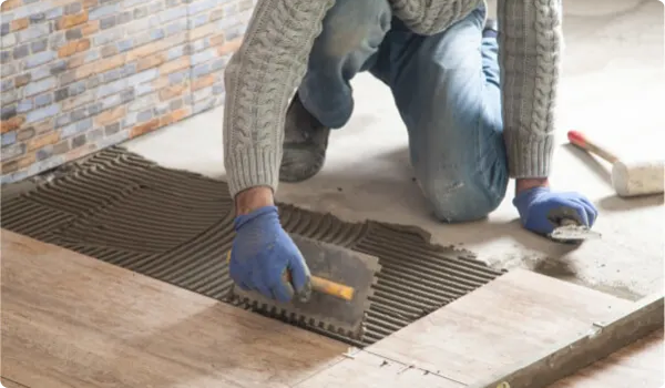 Person wearing blue gloves and a sweater kneeling while spreading adhesive with a notched trowel on a floor before laying wooden tiles.