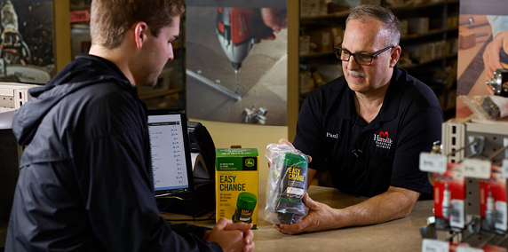 Store employee wearing glasses and a black shirt explaining a product to a customer seated at a counter in a hardware store with tools and product displays in the background.