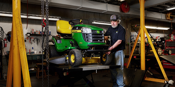 Man wearing a cap and casual work clothes inspecting a green and yellow lawn tractor lifted on a hydraulic platform inside a garage workshop.