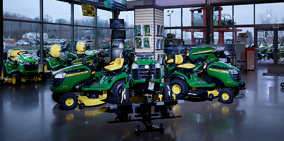 Indoor showroom featuring several green and yellow lawn tractors arranged on a polished concrete floor with large windows revealing an overcast outdoor scene.