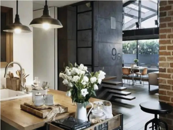 Cozy industrial-style kitchen and dining area with pendant lighting, wood countertops, white flowers in a vase, and large windows letting in natural light.
