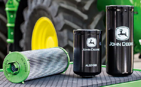 Three different John Deere branded filters in black and green, displayed on a textured surface with a large tractor tire blurred in the background.