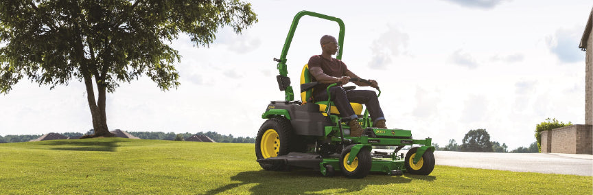Person operating a green and yellow zero-turn lawn mower on a large grassy lawn with a tree and house visible in the background on a partly cloudy day.