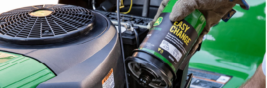 Close-up of a gloved hand pouring engine oil into a green riding mower, focusing on maintenance and home improvement tasks.