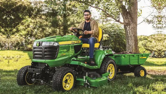Man wearing glasses and casual clothes driving a green riding mower with an attached trailer on a grassy lawn near trees in daylight