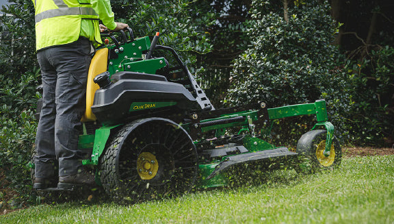 Person standing on a green and yellow stand-on mower cutting grass in a shaded garden area with dense green foliage in the background