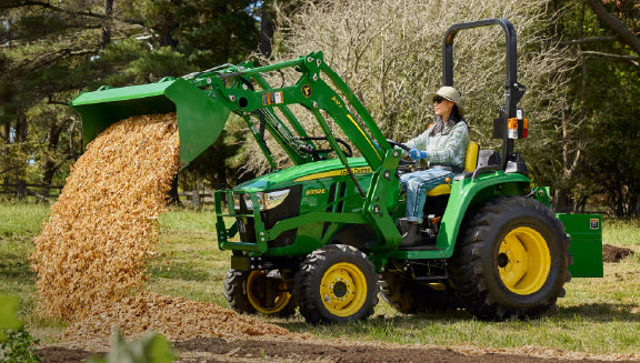 Person wearing a hat operates a green tractor with yellow wheels to dump a load of wood chips in a wooded outdoor area during daytime