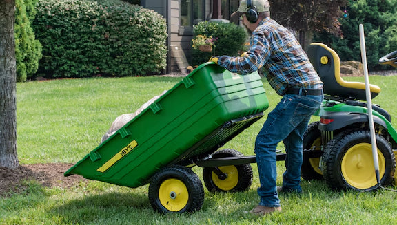 Person wearing ear protection and plaid shirt unloading a green utility cart attached to a green and yellow lawn tractor on a grassy lawn near a home