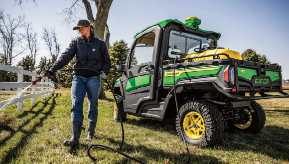 Person wearing a cap and jacket pressure washes a white wooden fence using equipment connected to a green off-road utility vehicle on a sunny day.